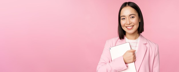 Image d'une jeune femme d'entreprise coréenne chef de la direction tenant une tablette numérique souriante et regardant un costume professionnel debout sur fond rose