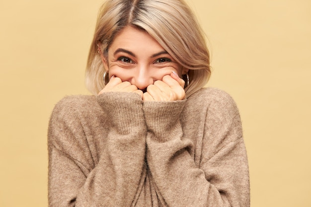 Image d'une jeune femme européenne joyeuse et joyeuse ne peut pas cacher ses émotions extatiques, être de bonne humeur, submergée par de bonnes nouvelles positives, se cachant la bouche derrière les mains, portant un pull confortable à manches longues