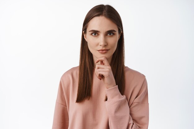 Image d'une jeune femme intelligente pensant, touchant le menton et ayant l'air heureuse et déterminée, réfléchissant à quelque chose d'intéressant, étant intriguée et souriante, fond blanc