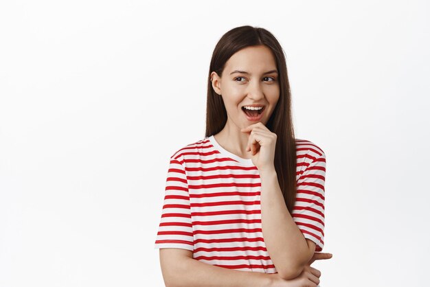 Image d'une jeune femme intriguée a une idée d'avoir l'air rusé et souriant, satisfait d'avoir l'air pensif sur le côté gauche en pensant avoir un plan debout sur fond blanc