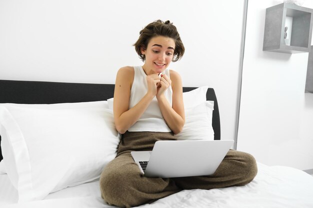 Photo gratuite image d'une jeune femme mignonne qui regarde avec espoir l'écran de son ordinateur portable en attendant quelque chose assis sur le lit