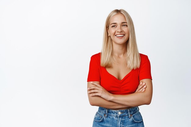 Image d'une jeune femme moderne détendue aux cheveux blonds, bras croisés sur la poitrine, souriante et regardant amicalement sur fond blanc de la caméra