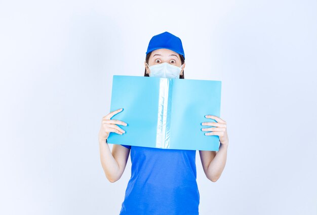 Image d'une jeune femme portant un masque médical posant avec un dossier bleu sur un mur blanc.