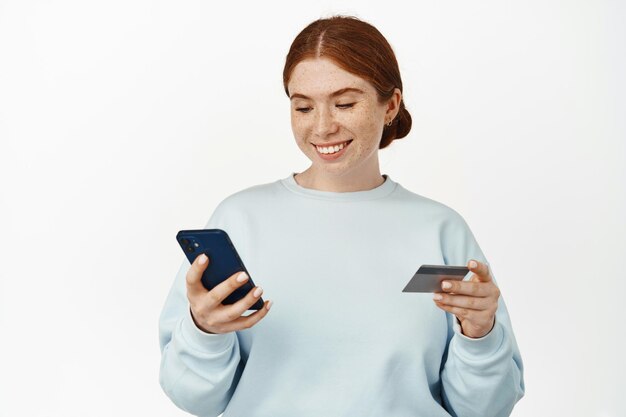 Image d'une jeune femme rousse souriante inscrivant sa carte de crédit dans l'application, regardant l'écran du téléphone portable, tenant une carte de réduction, debout sur fond blanc.