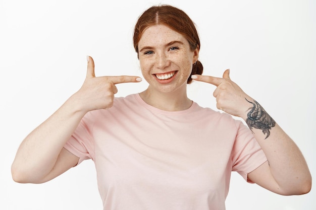 Image d'une jeune femme rousse souriante montrant son sourire blanc parfait, pointant vers des dents blanchies sans accolades, recommandant la stomatologie, la publicité du dentiste, fond blanc.