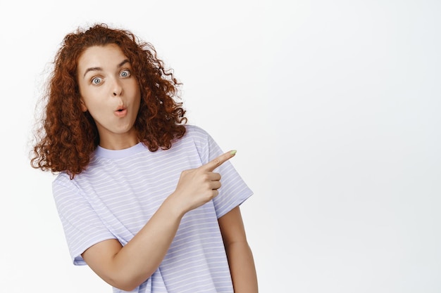 Image d'une jeune femme surprise pointant vers le coin supérieur droit. Fille aux cheveux rouges bouclés montrant la publicité de vente, le logo de l'entreprise, portant un t-shirt décontracté sur fond blanc