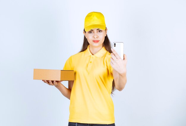 Image d'une jeune femme tenant un paquet et prenant un selfie sur un mur blanc.