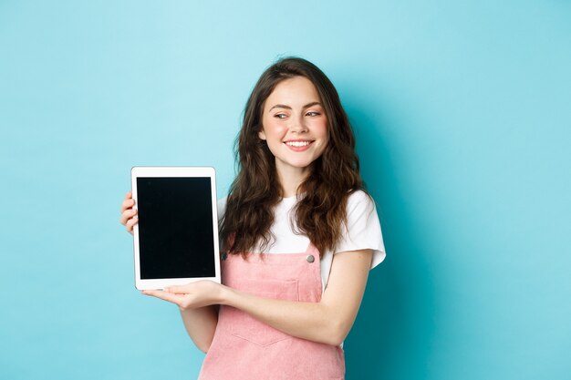 Image d'une jeune fille heureuse montrant un écran de tablette numérique et souriante fièrement, montrant votre logo à l'écran, debout sur fond bleu.