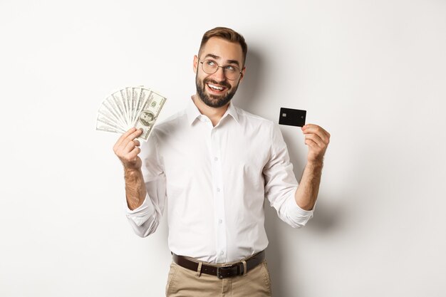 Image de jeune homme d'affaires détenant une carte de crédit et de l'argent, regardant le coin supérieur gauche et pensant au shopping, debout