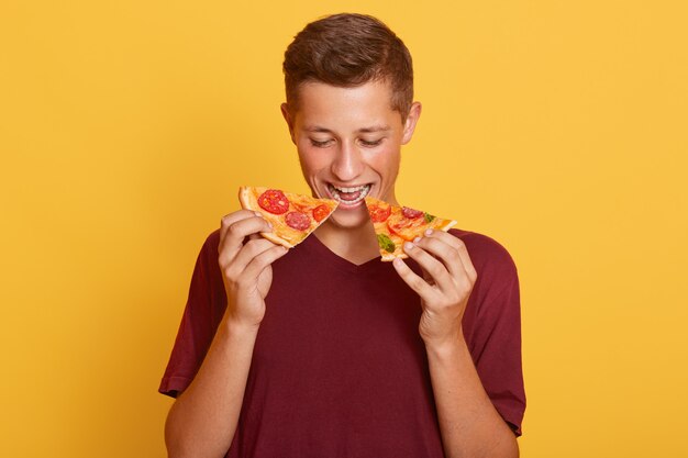Image de jeune homme affamé ravi de manger deux tranches de pizza, aimant la malbouffe, portant un t-shirt rouge, appréciant la nourriture savoureuse