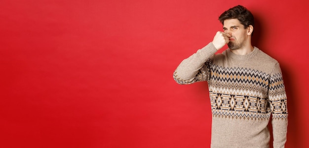 Image d'un jeune homme dégoûté en pull d'hiver fermé le nez et grimaçant de mauvaise odeur debout sur fond rouge