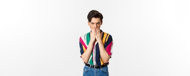 Image d'un jeune homme gay qui a l'air intrigué, tenant les mains sur la bouche, bavardant, debout sur fond blanc