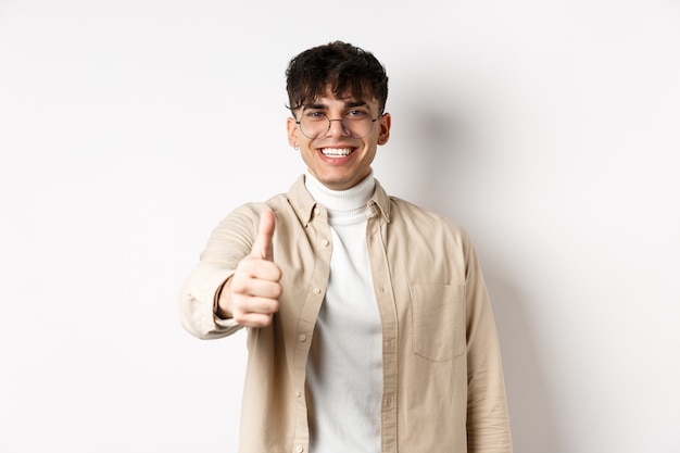 Image d'un jeune homme heureux montrant le pouce vers le haut et souriant satisfait, loue le bon travail, dis bien fait et a l'air heureux, debout sur fond blanc.