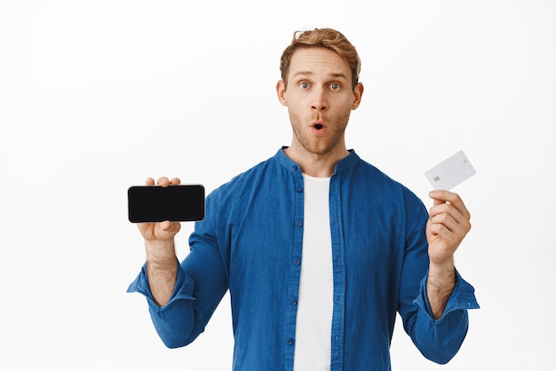 Image d'un jeune homme impressionné montrant un écran de téléphone mobile horizontal et une carte de crédit haletant et disant wow nouvelle banque propose une application pour de l'argent debout sur fond blanc