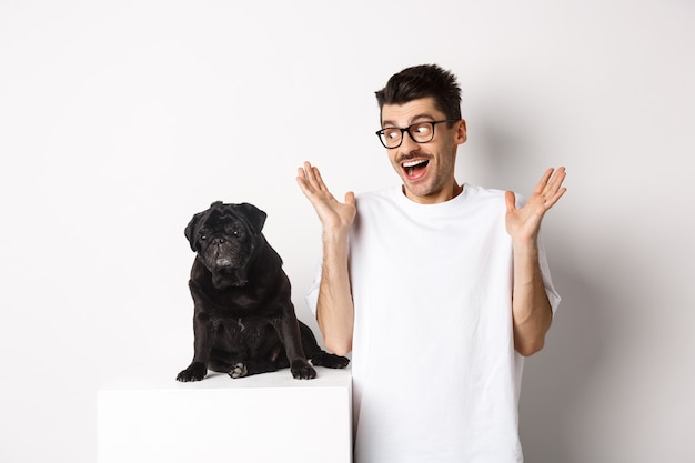 Image d'un jeune homme joyeux se réjouissant, regardant un mignon chien noir carlin et souriant, debout sur fond blanc.