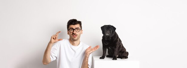 Image d'un jeune homme perplexe assis près d'un pug noir mignon montrant une petite taille et haussant les épaules confus