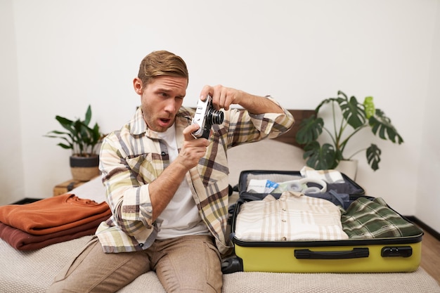 Photo gratuite image d'un jeune homme touriste vérifiant son appareil photo assis sur le lit avec la valise ouverte en train de photographier