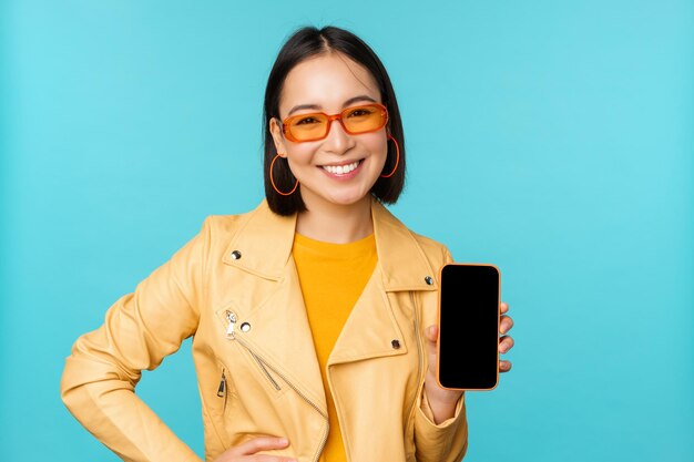 Image d'une jeune mannequin asiatique élégante portant des lunettes de soleil à la mode montrant l'application d'écran de téléphone portable sur l'interface du smartphone debout sur fond bleu