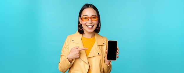 Image de jeune modèle féminin asiatique élégant dans des lunettes de soleil à la mode montrant l'application d'écran de téléphone portable