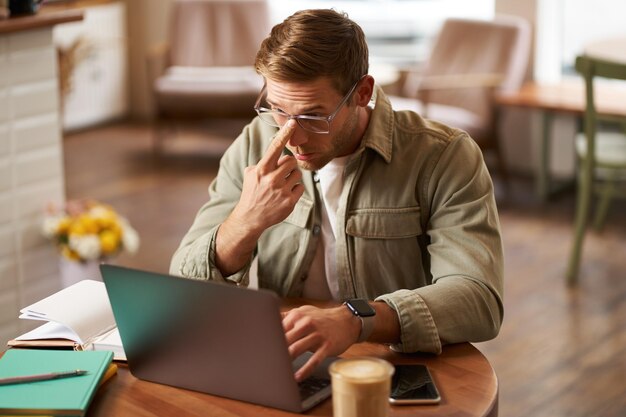 Image d'un jeune nomade numérique en lunettes assis dans un café qui travaille dans un café et utilise un ordinateur portable.