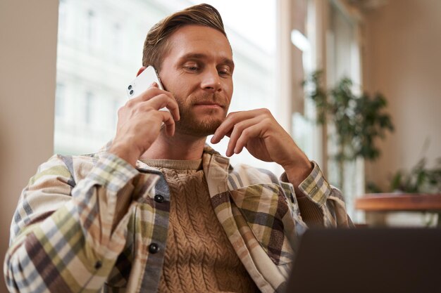 Image d'un jeune visiteur de café professionnel assis avec un ordinateur portable et parlant au téléphone portable à