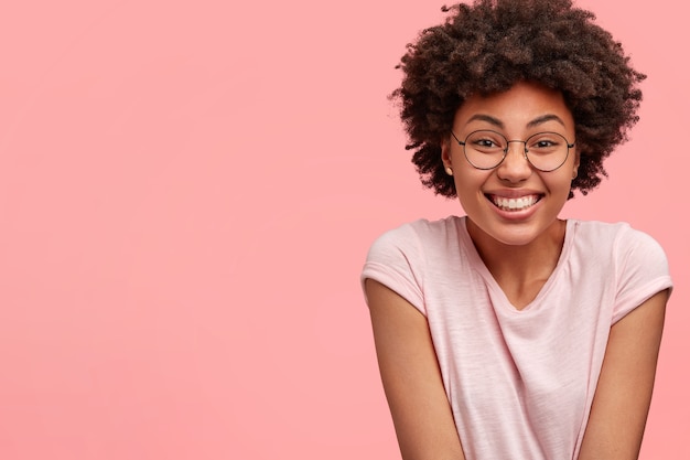 Photo gratuite image de jolie femme afro-américaine bouclée avec un sourire à pleines dents, a une expression joyeuse, se réjouit des moments agréables de la vie, vêtue d'un t-shirt décontracté, pose contre le mur rose avec espace de copie