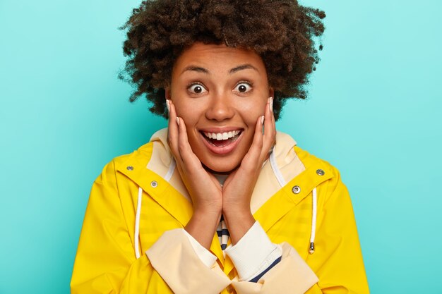 Image de jolie femme frisée avec une expression heureuse surprise, porte un imperméable jaune, sourit largement, a les yeux grands ouverts de l'émerveillement,