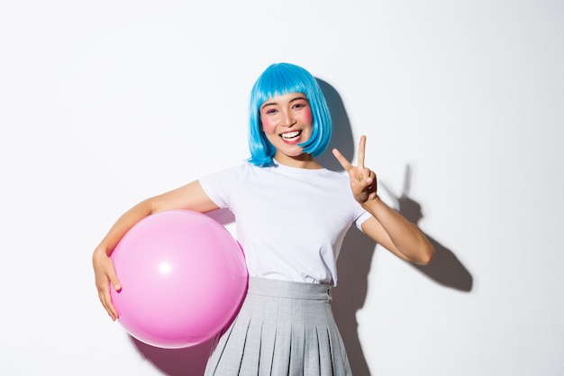 Image de jolie fille asiatique en perruque bleue et costume d'halloween, montrant le geste de paix, tenant un grand ballon rose.