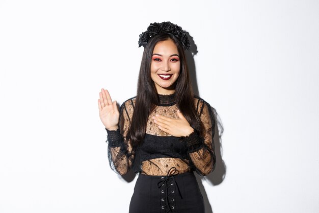 Image de jolie fille asiatique sincère en costume d'halloween faisant la promesse, tenant une main sur le cœur tout en jurant, être honnête, debout sur fond blanc.