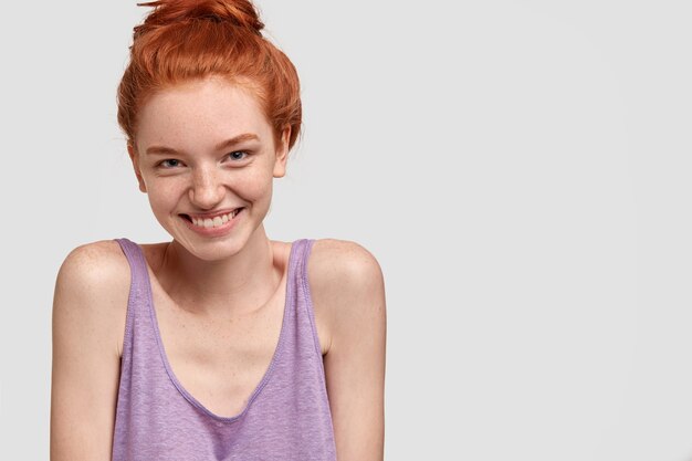 Image d'une jolie fille au gingembre avec un doux sourire à pleines dents, pose contre le mur blanc, vêtue d'un t-shirt décontracté, modèles d'intérieur, profite pleinement de la vie. Concept de féminité et de bonheur