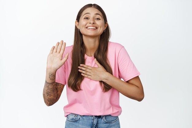 Image d'une jolie fille souriante mettant la paume sur le cœur et levant une main, faisant une promesse, se présentant ou faisant du bénévolat, debout sur fond blanc