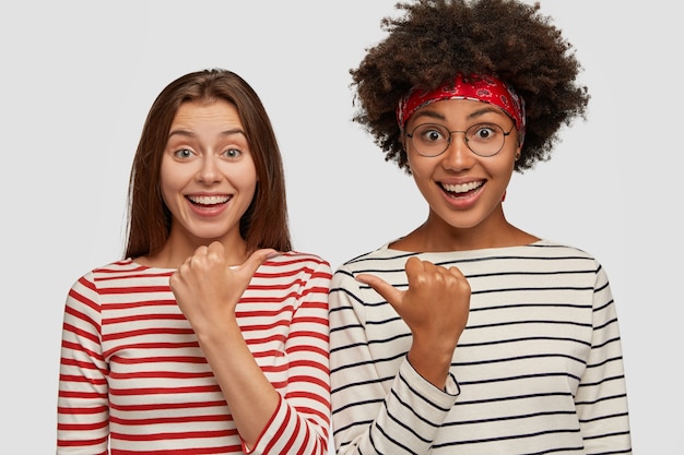 Image de jolies femmes avec des expressions joyeuses, sourire largement, se pointer du doigt avec les pouces, se sentir ravies, porter des vêtements rayés, modèle contre un mur blanc. Regardez mon ami, s'il vous plaît!