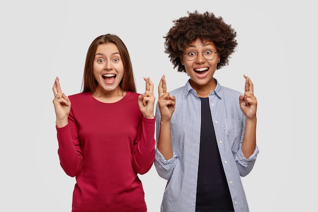 Image de joyeuses femmes multiethniques garder les mains levées avec les doigts croisés