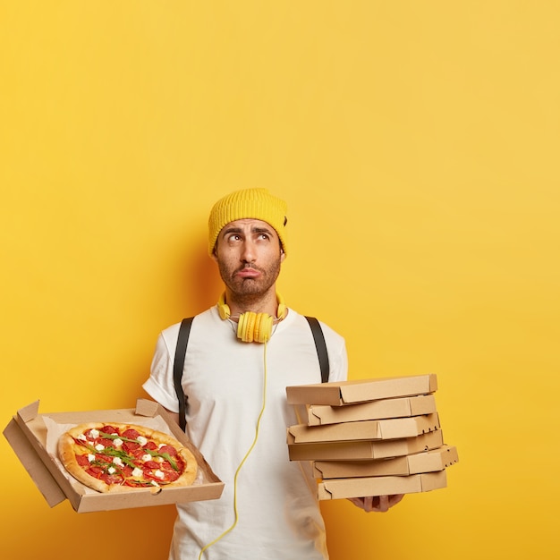 Image d'un livreur mécontent tient une pile de boîtes en carton, montre une savoureuse pizza au fromage, a une expression triste, porte un chapeau jaune et un t-shirt blanc