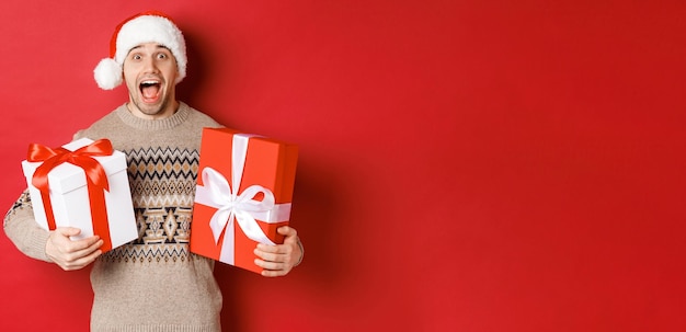 Image d'un mec attrayant et joyeux tenant des cadeaux de Noël, debout en bonnet de noel et pull d'hiver, souriant étonné, debout sur fond rouge.