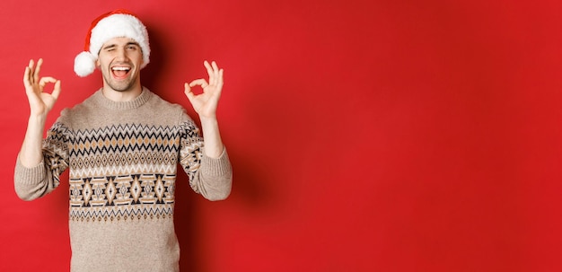 Image d'un mec séduisant et joyeux en pull et bonnet de Noel, souhaitant un joyeux Noël, montrant des signes d'accord et faisant un clin d'œil à la caméra, célébrant le nouvel an, debout sur fond rouge