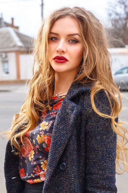 Image de mode en plein air de la belle femme élégante avec de longs cheveux blonds bouclés et de grandes lèvres pleines lumineuses posant dans la rue portant un manteau chaud élégant. Portrait d'automne.