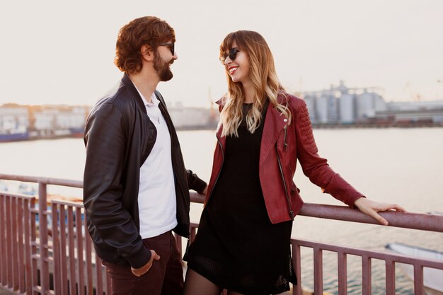 Image de mode en plein air d'un couple élégant en tenue décontractée, veste en cuir et lunettes de soleil debout sur le pont. Bel homme avec barbe avec sa petite amie, passer du temps romantique ensemble.