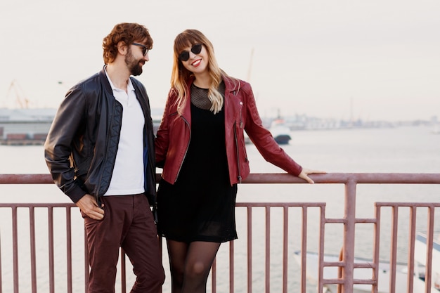 Image de mode en plein air d'un couple élégant en tenue décontractée, veste en cuir et lunettes de soleil debout sur le pont. Bel homme avec barbe avec sa petite amie, passer du temps romantique ensemble.