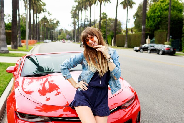 Image de mode de vie d'une femme de voyage assise sur le capot d'une incroyable voiture de sport décapotable rouge. Rues de Los Angeles