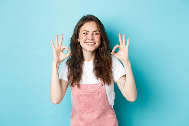 Image d'un modèle féminin brune souriante disant oui, montrant des signes d'approbation, d'accord ou faisant l'éloge d'un bon choix, recommandant un produit, debout sur fond bleu.