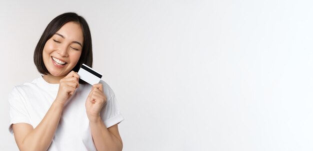 Image of smiling asian woman hugging credit card achat sans contact debout en tshirt blanc sur wh