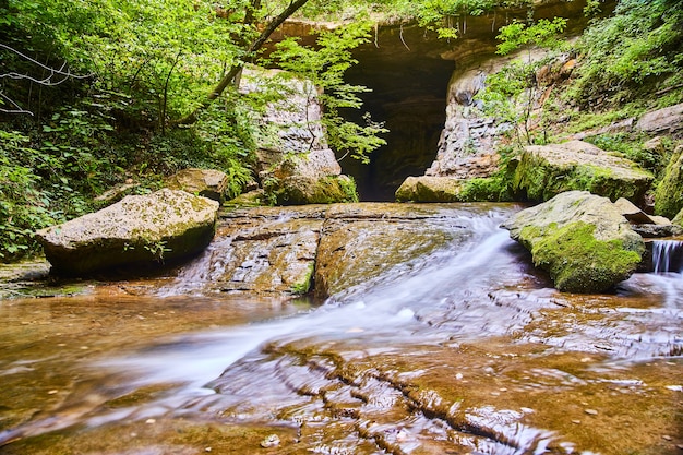 Image de l'ouverture d'une grotte à l'intérieur d'une forêt avec des arbres verts et une rivière blanche avec de gros rochers