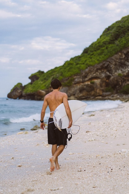 Image de paysage d'un surfeur masculin occupé à marcher sur la plage au lever du soleil tout en portant sa planche de surf sous son bras avec les vagues de l'océan se brisant en arrière-plan. Jeune beau surfeur masculin sur l'océan