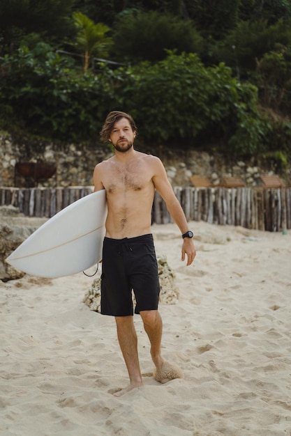 Image de paysage d'un surfeur masculin occupé à marcher sur la plage au lever du soleil tout en portant sa planche de surf sous son bras avec les vagues de l'océan se brisant en arrière-plan. Jeune beau surfeur masculin sur l'océan