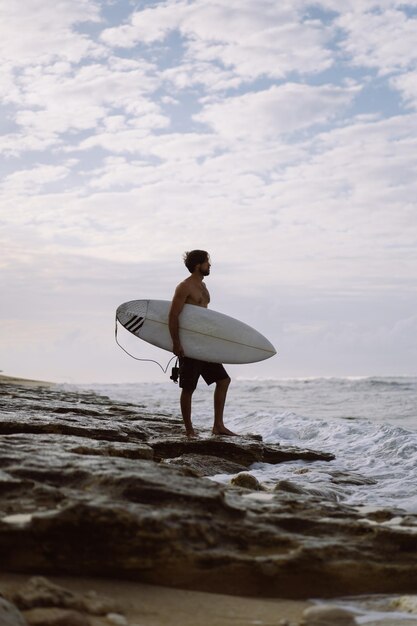 Image de paysage d'un surfeur masculin occupé à marcher sur la plage au lever du soleil tout en portant sa planche de surf sous son bras avec les vagues de l'océan se brisant en arrière-plan. Jeune beau surfeur masculin sur l'océan