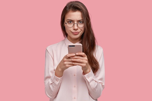 Image d'une petite amie sérieuse avec une expression faciale mécontente, presse les lèvres, porte un téléphone portable moderne, communique dans les réseaux sociaux