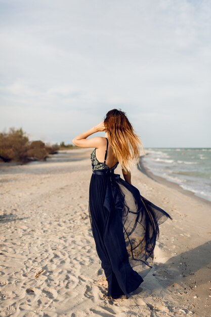 Image pleine hauteur de femme à la mode en robe de luxe élégante posant sur la plage. Vue de dos. Poils longs.