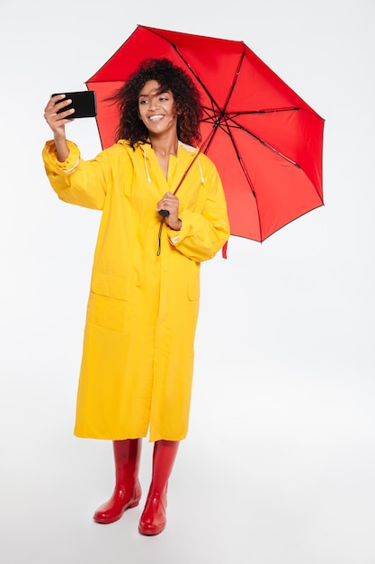 Image pleine longueur de femme africaine heureuse en imperméable se cachant sous un parapluie et faisant selfie sur son smartphone sur fond blanc