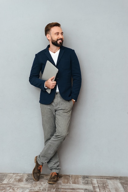 Image pleine longueur d'homme barbu heureux dans des vêtements de travail tenant un ordinateur portable et regardant loin sur gris